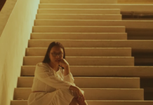 Shelma Shalindri Makes The Mundane Inspirational Indonesian musical artist Shelma Shalindri wearing a white dress and sitting at the bottom of a staircase with a soft, glowing light at the top of the staircase
