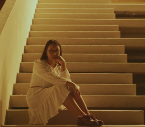 Indonesian musical artist Shelma Shalindri wearing a white dress and sitting at the bottom of a staircase with a soft, glowing light at the top of the staircase