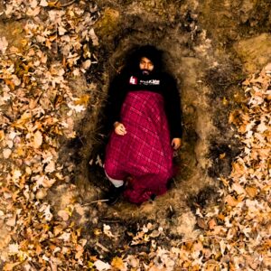 Zander, an American in a hole surrounded by autumn leaves wearing a hoodie and a blanket on top in his music video for the 2024 single "Know How To Die"