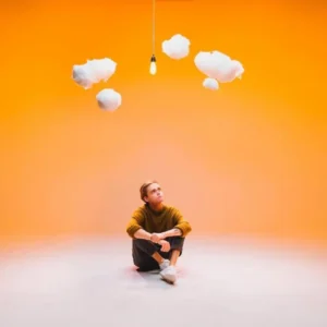 Indie singer-songwriter Devin Kennedy sitting beneath small, playful white clouds with an orange background as part of marketing material for his album "In My Imagination"