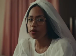 A close-up of singer-songwriter Jensen McRae sitting in a white wedding dress with a veil pulled back as she looks at the camera in the "Savannah" music video
