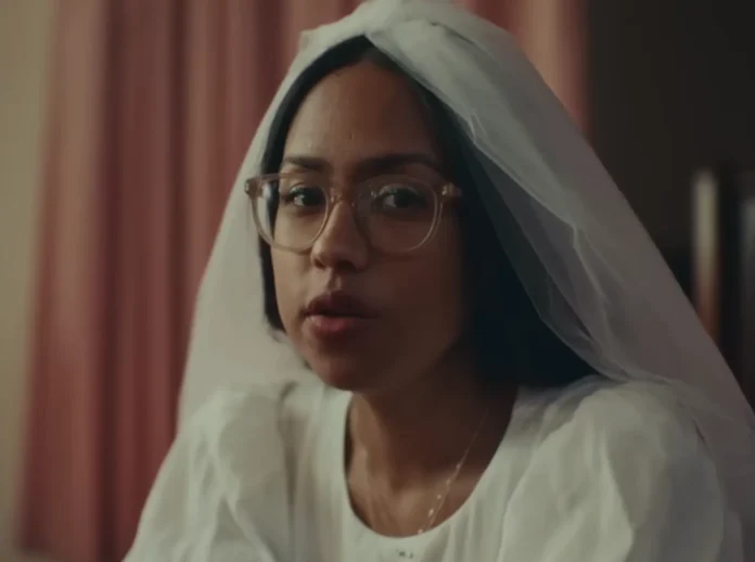 A close-up of singer-songwriter Jensen McRae sitting in a white wedding dress with a veil pulled back as she looks at the camera in the 