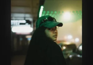 New Zealand alt R&B singer-songwriter JUGA pictured looking back at camera in a green hat with shades resting on top; cover art for "Late Night Talking"