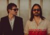 Brighton Duo Elandra Tear It All Apart in New Single “Ask Me Why” Brighton psychedelic pop-rock duo Elandra. (Left to right) bassist/co-producer George Seymour-Cole in a black blazer and shades; lead singer and co-producer Paolo Siviero in shades with shoulder-length brown hair and a patterned short-sleeved button-up shirt