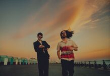 Elandra: A Conversation With The Promising Psych Rock Band Elandra band members George Seymour-Cole and Paolo Siviero in a suit and flowy patterned button-down shirt, respectively, with sunset-hued clouds and rows of houses on each side of them.