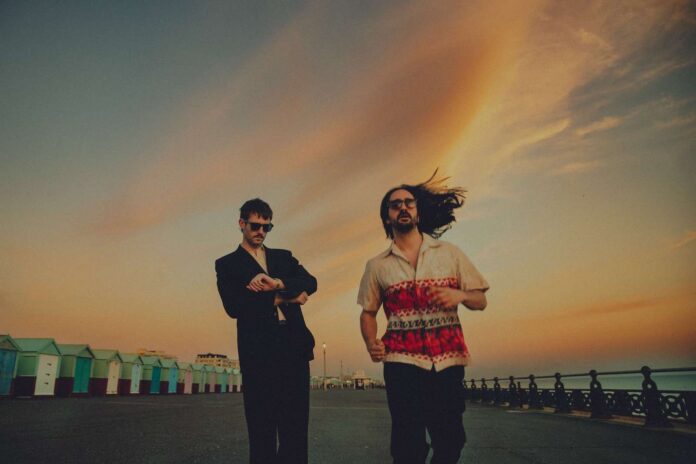 Elandra Band ATC Sound Interview Feature Photo Elandra band members George Seymour-Cole and Paolo Siviero in a suit and flowy patterned button-down shirt, respectively, with sunset-hued clouds and rows of houses on each side of them.