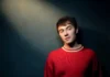 Sam Obrist on Making Music, Teaching Music, and Being an Out-and-Proud Late Bloomer NYC-based folk-pop artist Sam Obrist in a red sweater against a dark background with light shining down from the top left