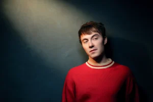 NYC-based folk-pop artist Sam Obrist in a red sweater against a dark background with light shining down from the top left
